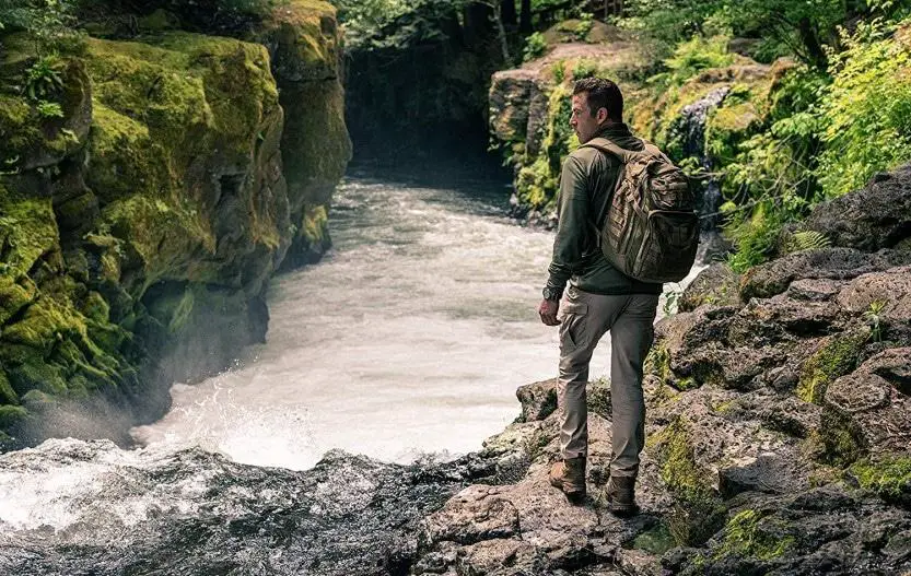 Hiker wearing a tactical backpack beside a fast-moving forest river